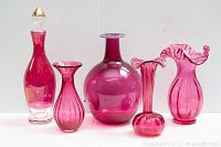 Front view of 5 cranberry glass items, including decanter with clear stopper and 4 vases of varying shapes and sizes on white background