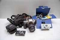 Wide shot showing Canon EOS Elan II camera with attached zoom lens, Panasonic Lumix camera in box, another compact camera, camera cases, and accessories on white background.