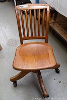 Full front view of vintage wooden office chair showing seat, back with vertical slats, and rolling casters on four legs.