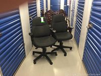 View of two black office chairs with armrests and caster wheels placed against a blue storage unit corridor wall showing their full shape and size.