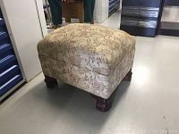 Square ottoman with floral upholstery and dark wooden carved legs on a concrete floor.