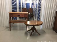 Two oak tables: a tall rectangular table with a drawer and a lower shelf, and a small round pedestal table. Shown in storage unit aisle with some wood chairs in background.