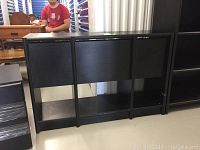 Black wooden tall bookshelf with three shelves on open bottom space, against white wall, on tiled floor