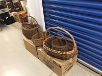 Four vintage wicker baskets of varying sizes and shapes placed on cardboard boxes. Natural brown wicker with some wear and patina.