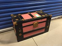 Front angle showing the chest's pink panels, black wood frame and metal corner embellishments, lock on front center.