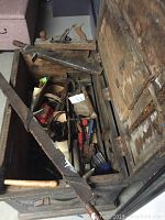 View inside wooden toolbox showing various hand tools including a large hand saw, hand planes and other tools.