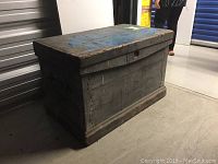 Side view of worn wooden chest with faded blue paint on top and metal corner reinforcements.