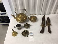 Full display of brass teapots, candlesticks, dinner bell, small cup and knives on white fabric surface.