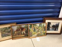 Four framed prints placed on floor against wall - three rustic house and garden scenes, one forest lake scene