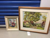 Photo showing both framed original oil paintings side by side on the floor against a blue storage unit door.