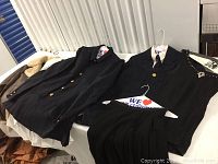 Photo of the man’s navy blue double-breasted suit jacket and lady’s black skirt and jacket with white blouse on hangers laid on table.