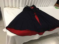 Dark navy blue wool nursing cape lying flat, showing clasp closure and red lining edges visible at bottom.
