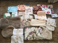 Flat lay of assorted linens including bathroom rugs, tablecloths, and fabric textiles in a variety of colors and patterns spread on carpeted floor