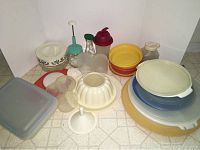 Collection of assorted Tupperware items including large bowls, lids, a bundt cake mold, and various plastic containers arranged on a tiled floor.