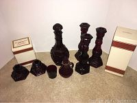 Full view of the Avon 1876 Cape Cod Decanter, five candlesticks, and two small cups on a beige carpeted floor, showing rich ruby red glass with textured pattern.