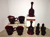 Complete set of Avon 1876 Cape Cod red glass items including creamers, sugar bowls, bells, and salt and pepper shakers displayed on boxes and carpet.