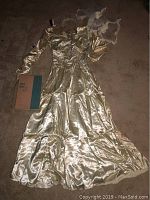 Full view of vintage satin wedding dress laid flat on floor with visible seedbead accents and lace-up back. Long sleeves visible, one torn. Vintage book beside dress titled 'Your Baby'.