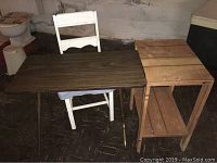 Photo of white painted wooden folding chair, rectangular wooden folding table, and small wood stand grouped together on dark floor.
