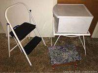 Photo showing white metal two-step stool, white metal mesh side table, plastic drawer container on top of the table, and a small floral cushioned foot stool on carpet.