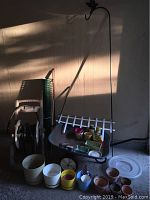 Wide shot showing rusty wheelbarrow filled with assorted small flowerpots, plant containers, garden chemicals, small spool of twine, and a white plastic trellis leaning against wall behind it. Also visible is a black metal shepherd's hook and a large green garden hose reel on the left as well as a white plastic garden stool.
