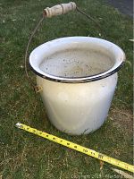 White enamel ware bucket with a metal wire handle and turned wooden grip, shown outdoors on grass with a measuring tape.