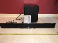 Samsung sound bar and wireless subwoofer displayed on floor with measuring tape showing about 40 inches length of sound bar, owner's manual placed in front of subwoofer.