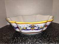 Large ceramic bowl with Italian decorative pattern on exterior, scalloped edges with yellow rim, placed on dark granite surface.