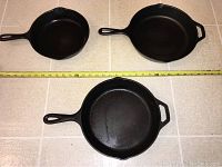 Three Lodge cast iron skillets with measuring tape showing sizes, two 12 inch and one smaller skillet.
