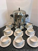Stainless steel coffee urn with eight white stoneware cups and saucers arranged on a countertop.