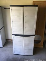Front view of the closed white plastic storage cabinet with black trim, showing the two doors with vented panel design.