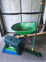 Photo showing the entire lot including the green wheelbarrow with wooden handles, green plastic garden stool, blue tarp folded underneath, and green push broom.