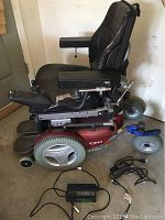 Left-side view of wheelchair showing maroon base, black seat, joystick control arm, footplate and charger cables on floor.