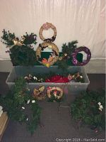 Overview of assortment of wreaths displayed above and inside clear storage bin, showing pine needle artificial wreaths decorated with bows, pine cones, and baubles.