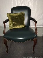 Front view of the green leather chair with the velvet pillow on the seat showing upholstery, wooden arms and legs, and nailhead trim.