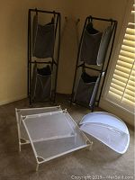 Ironing board, two double hampers on wheels and 4 shirt drying nets arranged on carpeted floor near window blinds.
