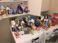 Photo showing various cleaning sprays, detergents, polishes, and household products on top of a washer and dryer set.