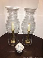 Two clear glass chimneys with brass candlesticks inside and white taper candles, along with a small white marble grinder placed in front of them on a wooden surface.