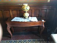 Wood console table with carved legs, decorative apron, and glass top shown against wood-paneled wall.