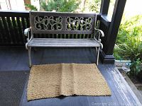Photo showing the entire cast iron park bench with decorative wood back slats and matching wood seat, positioned behind a woven outdoor carpet on a wooden porch floor.