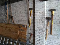 Photo showing two axes with wood and plastic handles, pickaxes, shovels, sledge hammer and other tools hanging on white brick wall.