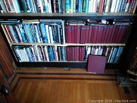 Bookshelf showing maroon hardcover postcard albums tightly packed on shelves next to a variety of other hardcover books mostly historical and nautical topics.