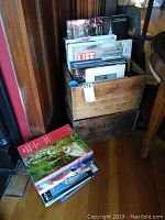 Two wooden crates stacked containing books and magazines with a couple of books on the floor next to them.