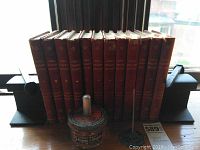 Photo of twelve red hardcover volumes of John L. Stoddard's Lectures standing upright between two dark metal railroad rail bookends with railroad spikes, on wooden surface near window.