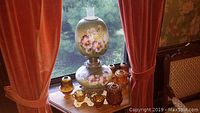 Photo showing an amber glass oil style electric lamp with floral hand-painted design surrounded by amber glassware pieces on a wooden table.