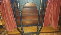Side view showing black turned legs and stretcher base of the wooden side table against wooden paneling and flanked by rust-colored curtains.