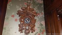 Front view of the wood cuckoo clock showing the detailed carved hunting motif with deer head, crossed rifles, oak leaves, black clock face with white Roman numerals.