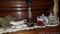 Side view of gravy boat, serving tray, candle holders, salt and pepper shakers, cupcake candle, and two ceramic teapots arranged on lace doily on wooden surface