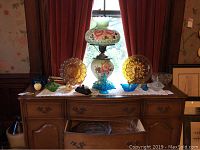 Full view of the dresser top showing the 8 pieces of decorative glassware in clear, blue, amber, and green with a pale green oil-style electric lamp with floral pattern and metal base in the center.