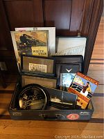 Photo of vintage suitcase filled with framed travel and nautical art including Queen Mary tin plate and posters