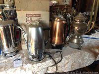 Photo showing five vintage percolators arranged on tablecloth, one in original Universal Coffee-matic box, varying materials including stainless steel, copper-tone and brass-tone.